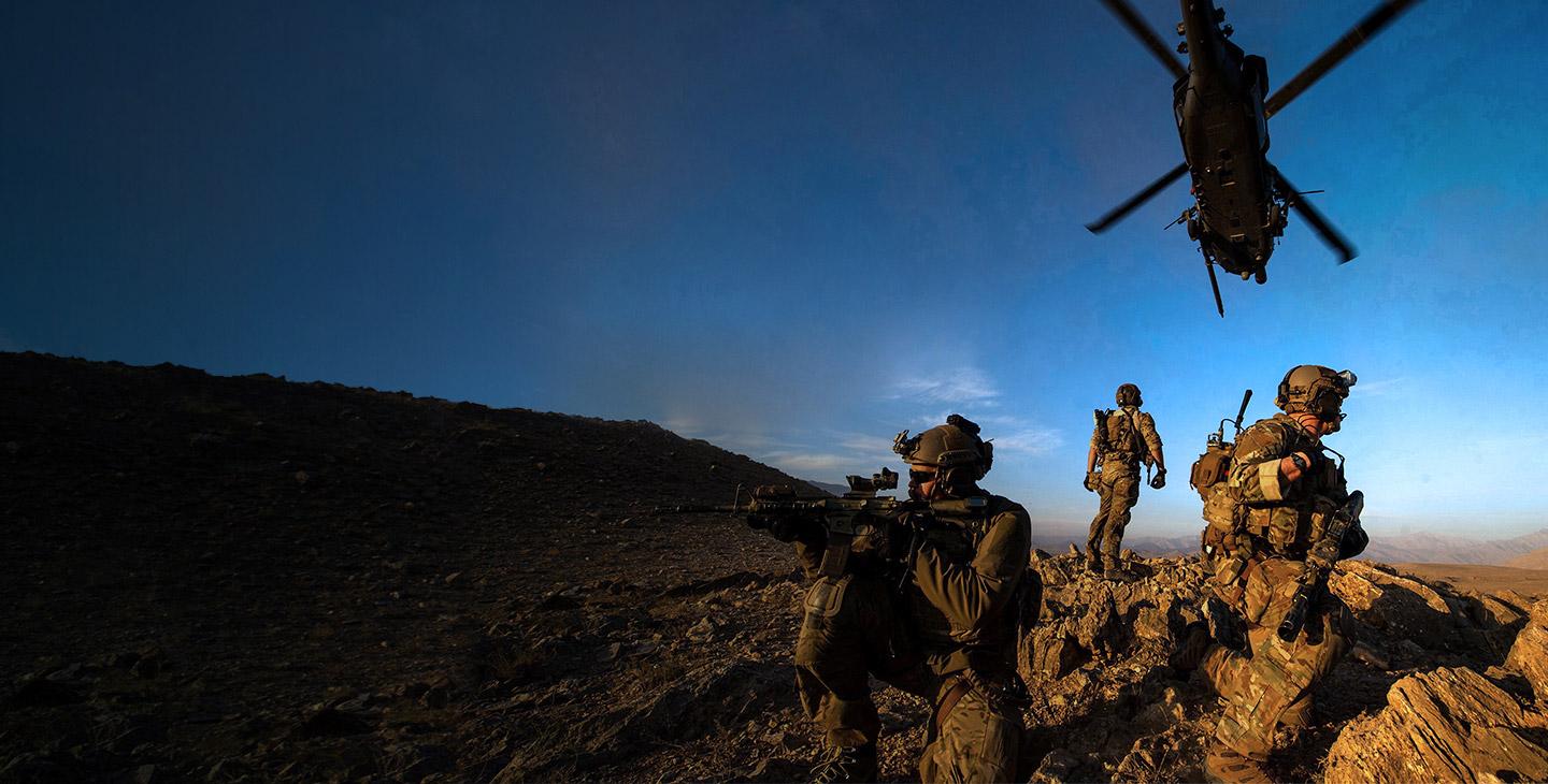 3 military men dressed in combat gear on watch on top of a rocky hill with a helicopter flying overhead